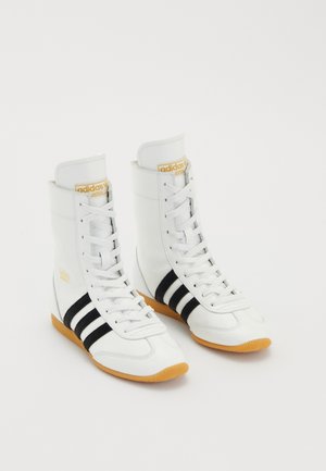 Witte hoge sneakers van leer, met drie zwarte strepen, witte veters en een gumrubberen zool. Gouden logo-accent op de tong.