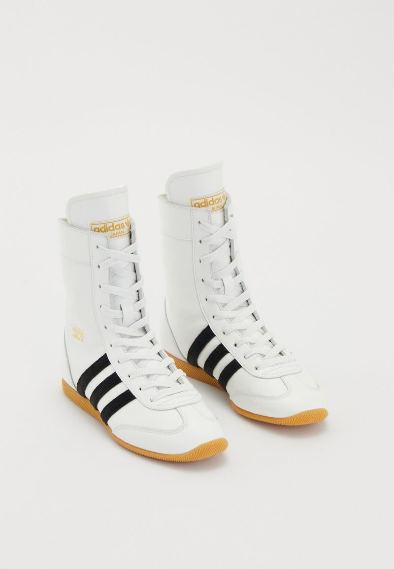 Weiße High-Top-Sneaker aus Leder, mit drei schwarzen Streifen, weißen Schnürsenkeln und einer Gummisohle. Goldenes Logo-Detail auf der Zunge.