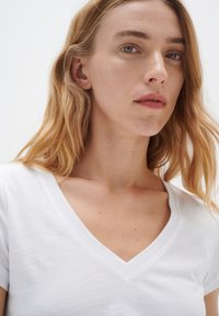 Bílá V- neck tričko s krátkými rukávy, hladká textura a minimalistický design, jasně zobrazuje výstřih na neutrálním pozadí.