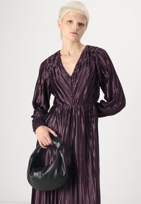 Raukšlėta tamsiai violetinė suknelė su ilgomis rankovėmis ir V formos iškirpte, derinama su juoda, glotnia tekstūra rankine, laikoma kairėje rankoje.