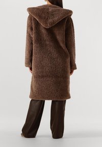 Bruine, fluffy capuchonjas met een gestructureerd oppervlak, lange mouwen en een knielange snit, gecombineerd met donkere wijde broekspijpen.