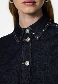 Mørk denimskjorte med knappnedkrage, metall klaffeknapper og dekorative sølvsømmer langs kragen med kontrasterende søm.