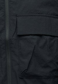 Κοντινό πλάνο μαύρου υφασμάτινου σακακιού που δείχνει ένα φερμουάρ και μια μεγάλη ορθογώνια τσέπη με καπάκι.