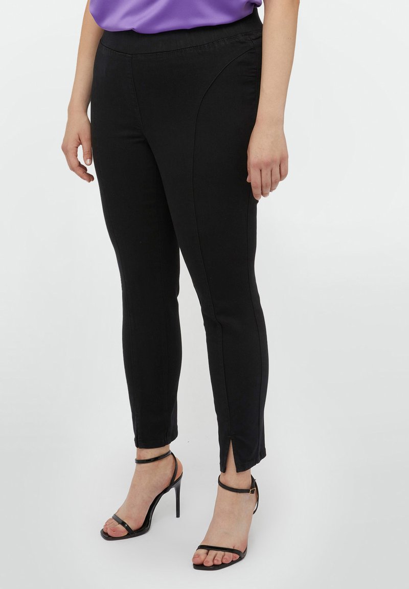 Pantalones negros de corte ajustado con aberturas laterales, hechos de un material elástico. Presentan una textura suave y un diseño de cintura alta, combinados con tacones atados.