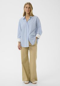 Chemise boutonnée à rayures bleu clair avec des poignets blancs, associée à un pantalon large couleur beige et des chaussures crème, présentée sur un fond uni.