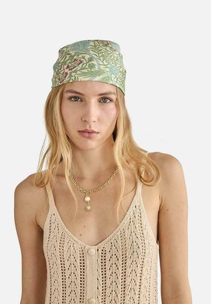 Joven mujer con cabello largo y rubio que lleva un pañuelo verde con flores, top beige sin mangas de punto y collar de cadena dorada con colgantes de sol y perla.