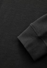 Tissu en tricot gaufré gris foncé avec un poignet lisse. La texture semble côtelée, soulignant la chaleur et un look décontracté. Lignes épurées, détails minimaux.