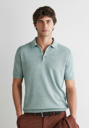 Mężczyzna o brązowych włosach ma na sobie jasnooliwkowy polo z krótkim rękawem wpuszczony w brązowe spodnie, stoi na jednolitym białym tle.