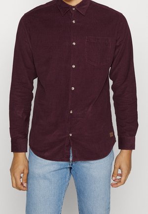 Man in een donker bordeauxkleurig corduroy overhemd met knopen en een borstzak, met lichtblauwe jeans, staande tegen een effen achtergrond.