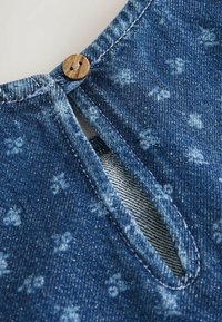 Jeans tkanina z modrim in belim cvetličnim vzorcem, z odprtino spredaj v obliki ključavnice, ki jo zapira leseni gumb. Vidna je teksturirana šivanka.