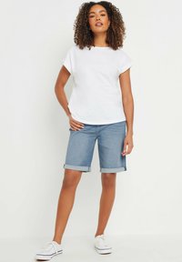 Camiseta blanca de manga corta con escote redondo, combinada con pantalones cortos de mezclilla de Bermuda en un tono azul claro con dobladillos enrollados y zapatillas blancas.