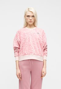 Νεαρή γυναίκα με μακριά ξανθά μαλλιά, που φοράει ροζ φούτερ με animal print και ταιριαστό ροζ παντελόνι, στέκεται μπροστά σε απλό λευκό φόντο.