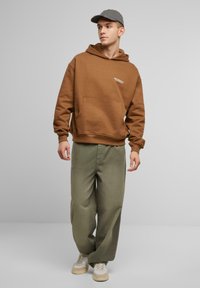 Brun hættejakke med en frontlomme og logodetalje, parret med løstsiddende olivengrønne bukser og lyse sneakers. Grå kasket fuldender looket.