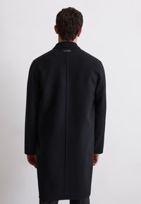 Langer schwarzer Wollmantel mit geradem Schnitt, einem Schlitz im Rücken und einem dezenten Markenlabel am Kragen. Glatte Textur, ohne Muster.