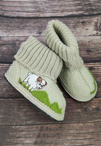Beige Wollpantoffeln mit gerippten Bündchen und einem aufgestickten Cartoon-Widder auf grünen Hügeln auf einem Pantoffel, liegend auf Holzdielen.