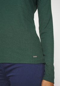 Gros plan sur une personne portant un haut à manches longues côtelé vert foncé et un pantalon bleu marine, montrant une petite étiquette métallique de marque près de l'ourlet.