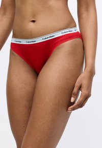 Piros pamut alsónadrág fehér elasztikus derékpánttal, amelyen fekete "Calvin Klein" logó található. Puha tapintás, alacsony derék, minimalista fazon.