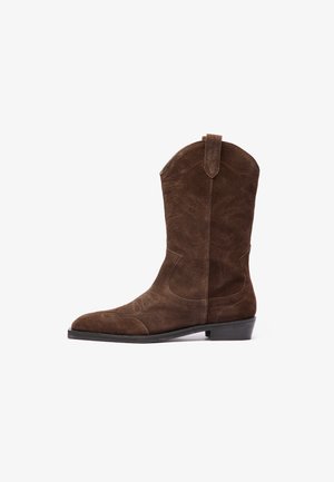 Bota de cowboy em camurça castanha com biqueira pontuda, detalhes de costura em relevo e salto baixo empilhado. Apresenta alças laterais para facilitar ao calçar.