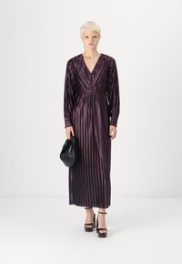 Violetinė plisuota maksi suknelė su V formos iškirpte, ilgomis rankovėmis ir pabrėžta juosmeniu. Pora su juodu rankiniu kosmetiku ir platforminėmis aukštakulnėmis.