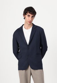 Marineblå strukturert blazer med to knapper, notched lapels og sidepokker, båret over en hvit skjorte og lyse beige bukser.