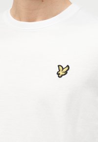 Άσπρο μπλουζάκι με στρογγυλή λαιμόκοψη που διαθέτει ένα μικρό κίτρινο πουλί με μαύρο περίγραμμα στο αριστερό μέρος του στήθους.