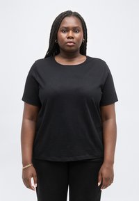 Γυναίκα με μακριά πλεγμένα μαλλιά, φορώντας ένα απλό μαύρο μπλουζάκι και μαύρο παντελόνι, στέκεται μπροστά σε ανοιχτό γκρι φόντο.