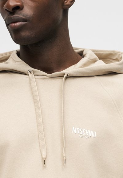 Gros plan sur une personne portant un sweat à capuche beige avec des cordons et le logo blanc "Moschino Active" sur la poitrine.