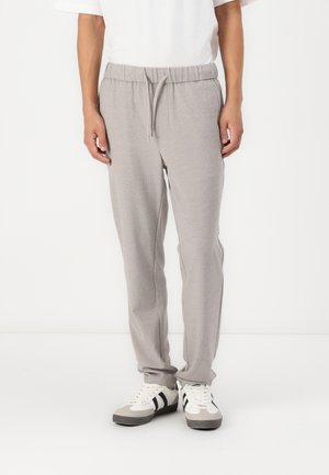 Person trägt hellgraue Jogginghosen mit Kordelzugbund und weiße Turnschuhe mit schwarzen Streifen, steht vor einem schlichten weißen Hintergrund.