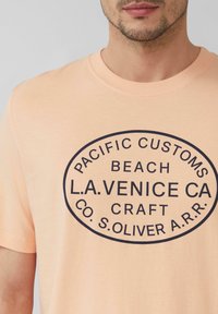 Perzikkleurig T-shirt van katoen, met een zwart ovale graphic met tekst. Crewneck ontwerp, korte mouwen en een gladde textuur.