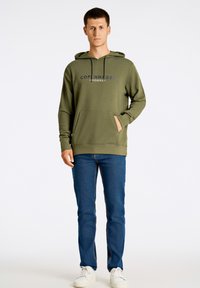 Sudadera color verde oliva con un acabado texturizado, bolsillo frontal y logo impreso "COPENHAGUE". Combinada con jeans de mezclilla azul y zapatillas blancas.