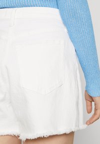 Pantalones cortos de mezclilla blanca con un dobladillo deshilachado, que cuentan con un único bolsillo trasero y un suéter azul texturizado parcialmente visible.