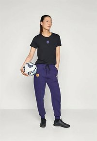 Camiseta negra con el logo del FC Barcelona, pantalones de chándal morados con logo y bolsillos con cremallera. Sosteniendo un balón de fútbol blanco y azul, lleva puestas unas zapatillas negras.