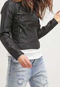 Zwarte jas van imitatieleer met een aangesloten ontwerp, ritszak aan de zijkant en subtiele stiksel details, gecombineerd met licht beschadigde spijkerbroeken.