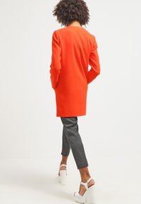 Světlý červený dlouhý blejzr se rovnými liniemi, s dvěma bočními kapsami, v kombinaci se šedými slim kalhotami a bílými sandály na vysokém podpatku.