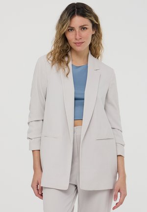 Blazer grigio chiaro con vestibilità morbida, rever a punta e due tasche frontali. Abbinato a un top blu aderente, che presenta una texture di tessuto liscia.