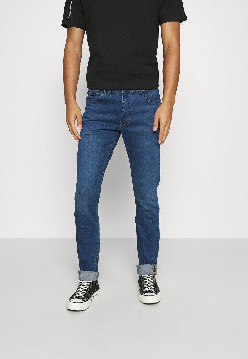 Man som bär slim-fit blå jeans med uppvikta fållar, svart T-shirt och svarta högklackade skor med vita snören, stående på ett grått golv.