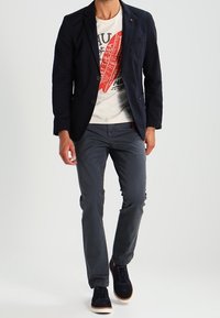 Ciemnoniebieska marynarka na białym t-shircie z grafiką i czerwoną nadrukiem, zestawiona z szarymi spodniami i ciemnymi sneakersami z białymi podeszwami.