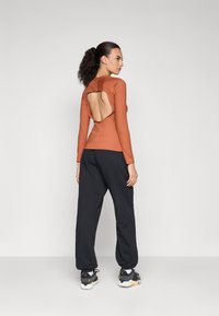 Langermet oransje topp med en stor åpen rygg, paret med sorte bukser med snøring. Antrekket har en glatt tekstur og en tettsittende design.