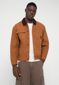 Giacca di jeans marrone con colletto in velluto a coste, bottoni a pressione frontali, due tasche sul petto e vestibilità rilassata, indossata sopra una camicia bianca e pantaloni marroni.