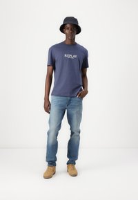 Camiseta azul marina con el logo blanco "REPLAY", combinada con jeans azul claros y zapatillas color beige, complementada con un sombrero de cubo oscuro.