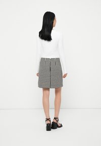 Kobieta z długimi czarnymi włosami ubrana jest w białą bluzkę z długim rękawem, spódnicę w czarno-białą kratkę houndstooth z zamkiem z tyłu oraz czarne sandały na wysokim obcasie.