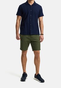 Camicia button-up a maniche corte blu navy con colletto, abbinata a pantaloni corto verde oliva. Il modello indossa sneakers blu navy. Outfit semplice e casual.