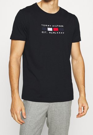 Uomo che indossa una t-shirt nera Tommy Hilfiger con logo e pantaloni grigi, in piedi con le braccia rilassate ai lati.