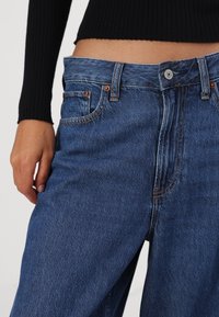 Modré džíny s vysokým pasem, obsahující měděné nýty, přední zapínání na knoflík a viditelné švy.