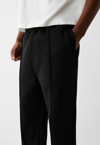 Black tapered pants with a smooth texture, elastic waistband, and side pockets. Worn with a white shirt, displaying a simple design.