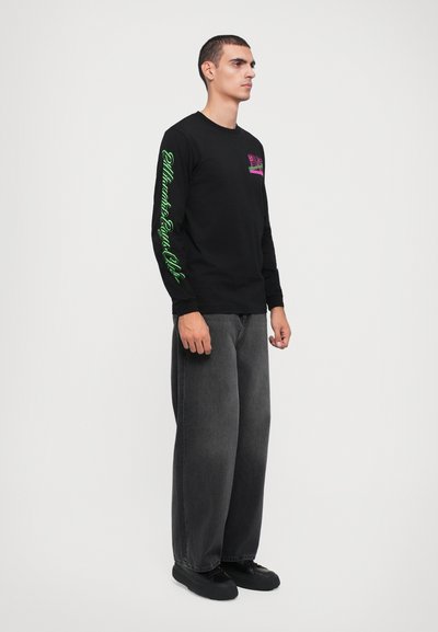 Camisa preta de manga longa com gráficos rosa e verde. Combinada com jeans escuros de corte largo e sapatos pretos. Design simples e casual.
