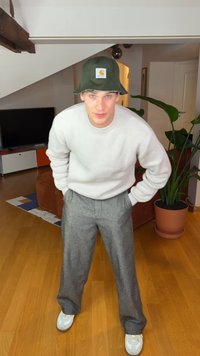 Hellgrauer, strukturierter Pullover, graue karierte Hose, grüner Bucket-Hut mit Logo-Patch, weiße Turnschuhe. Innenraum mit Pflanzen und Möbeln.