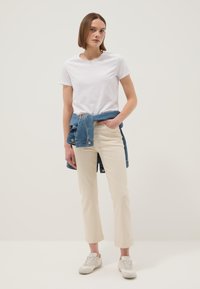 Mujer de pie con la mano en el bolsillo, vistiendo una camiseta blanca, pantalones beige, zapatillas blancas y una chaqueta vaquera atada alrededor de la cintura.