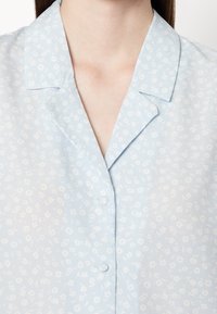 Light blue blouse featuring a subtle floral pattern, short collar, and small matching buttons along the front. Soft fabric texture.