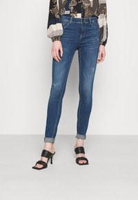 Jeans ajustados de denim azul con un acabado desgastado, puños enrollados y bolsillos frontales, combinados con sandalias de tacón alto negras.